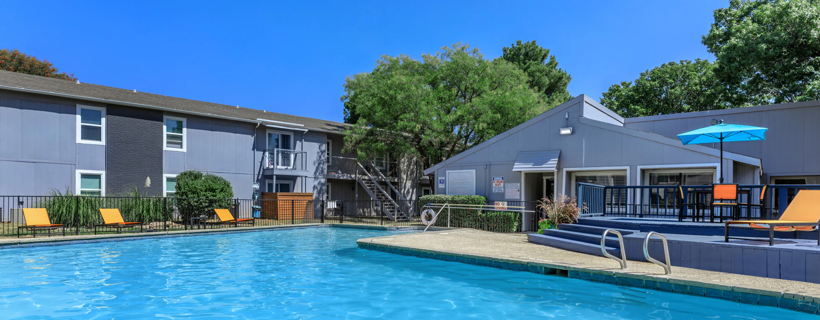 Bright, modern apartment community featuring a large swimming pool, sun loungers, and a clubhouse with a shaded patio area, surrounded by two-story gray buildings and mature trees.