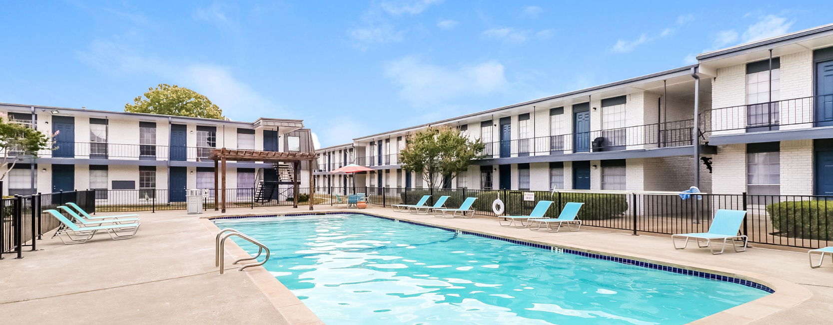 Classic two-story apartment complex with a central swimming pool, surrounded by white brick buildings with blue doors and trim, and turquoise lounge chairs lining the pool deck.