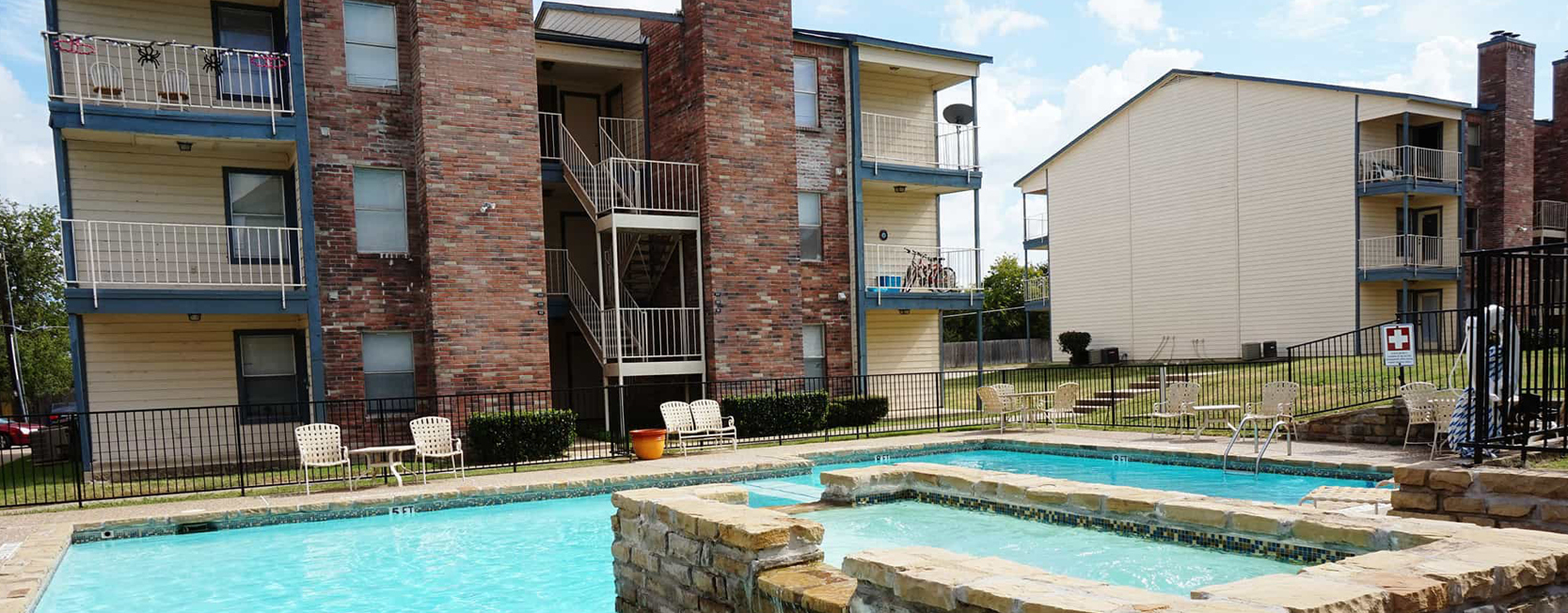 Three-story brick and siding apartment complex with central outdoor pool, stone hot tub feature, and fenced-in seating area under a bright blue sky.