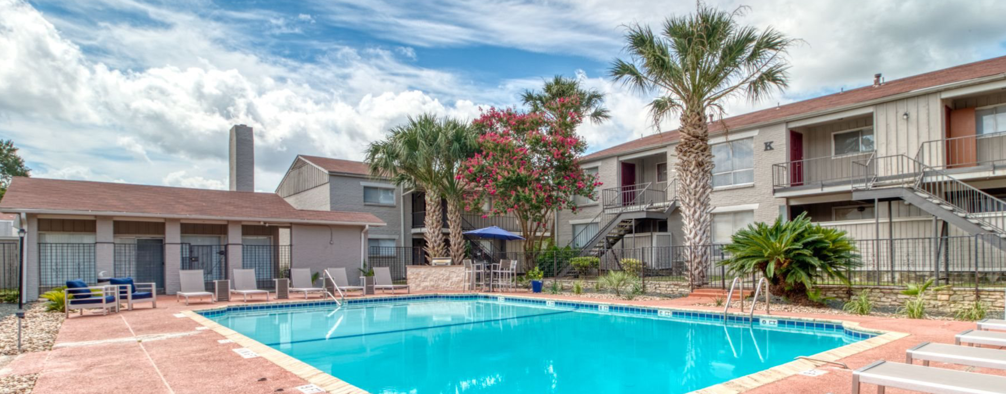 The Ascent Apartments, two-story apartment building, with a swimming pool in the foreground surrounded by palm tree and seating.