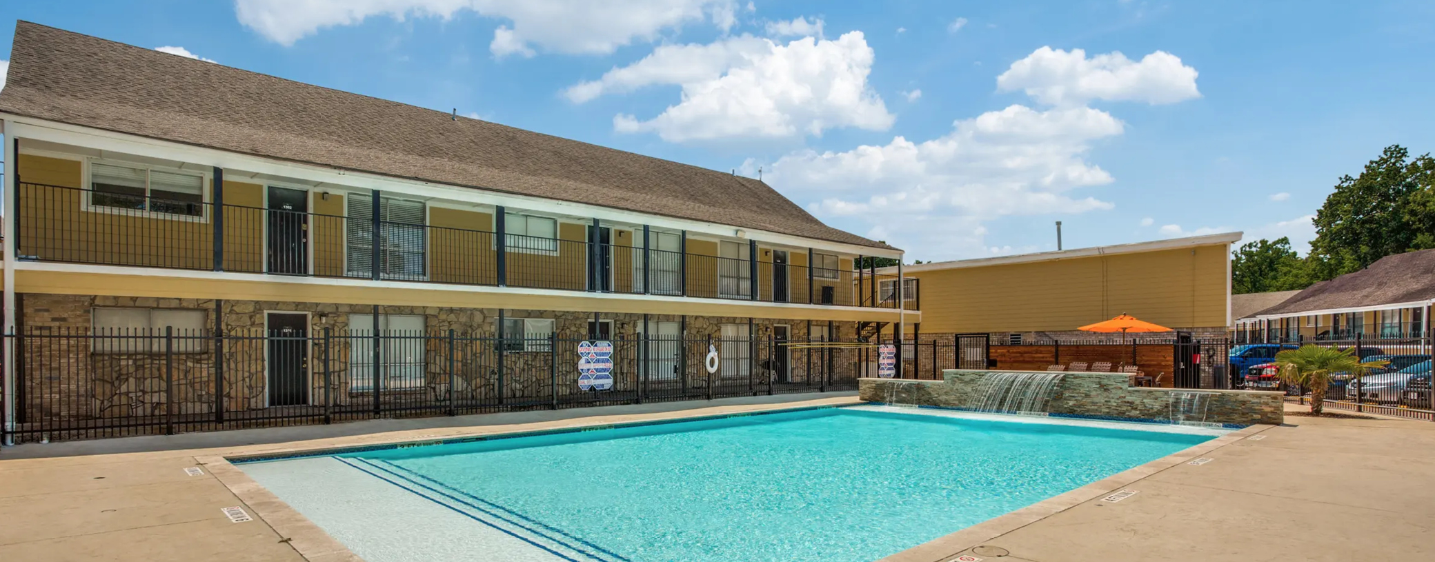 Two-story, beige brick, apartment building with a sparkling pool in the foreground during a partly cloudy blue sky day.