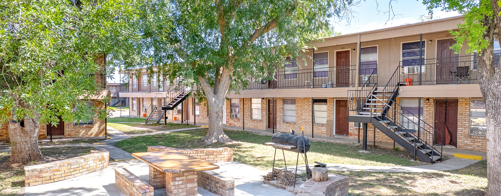 Two-story brick apartment complex with exterior staircases, brown doors, shaded courtyard with mature trees, and a central outdoor BBQ area with built-in brick benches and picnic tables.