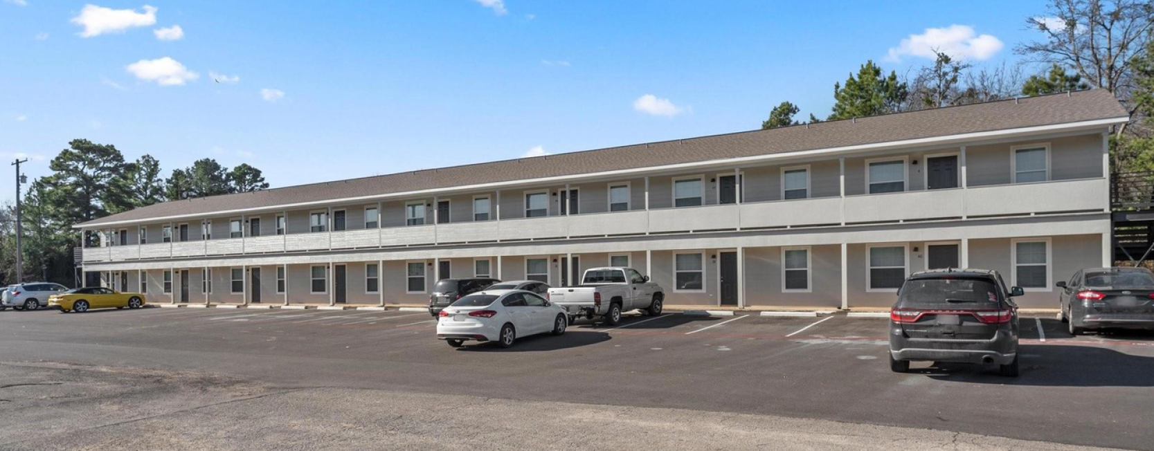 A two-story, motel-style apartment building with a long balcony and parking lot in front, surrounded by trees and several parked vehicles under a clear blue sky.