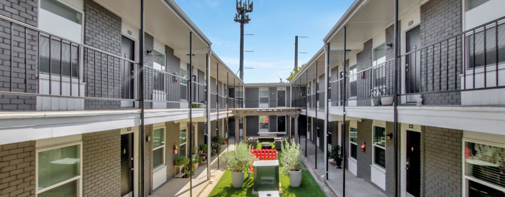 Modern two-story apartment complex with gray brick exterior, black doors, metal railings, and a courtyard featuring potted plants and oversized Connect Four game.