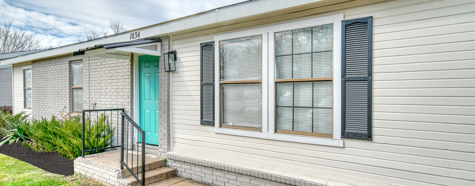 A charming brick-and-siding single-story home with black shutters, large front windows, and a turquoise door framed by shrubs and a short staircase.