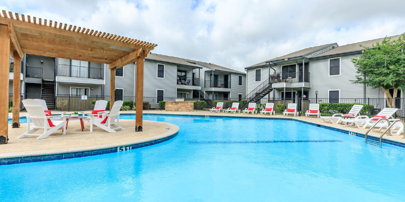 A modern apartment complex features a large outdoor pool surrounded by white lounge chairs with red pillows, a shaded pergola seating area, and two-story buildings with gray siding and black metal staircases.