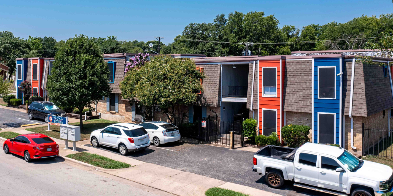 Two-story apartment complex with colorful orange and blue accents, featuring sloped roofs, and a parking area with several vehicles in front.