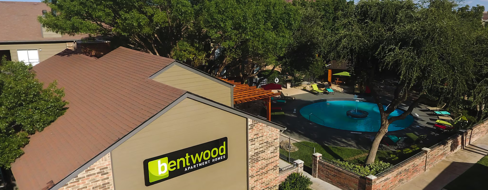 Aerial view of an apartment community with a landscaped courtyard, swimming pool, mature trees, and a building displaying an apartment complex sign.