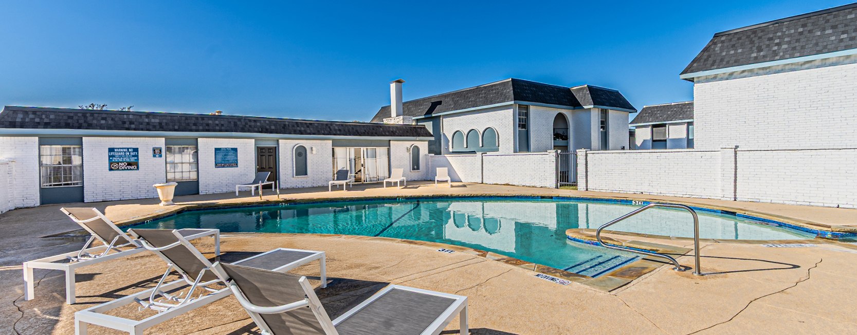 Outdoor swimming pool area at an apartment community with white brick buildings, lounge chairs, and clear blue sky.
