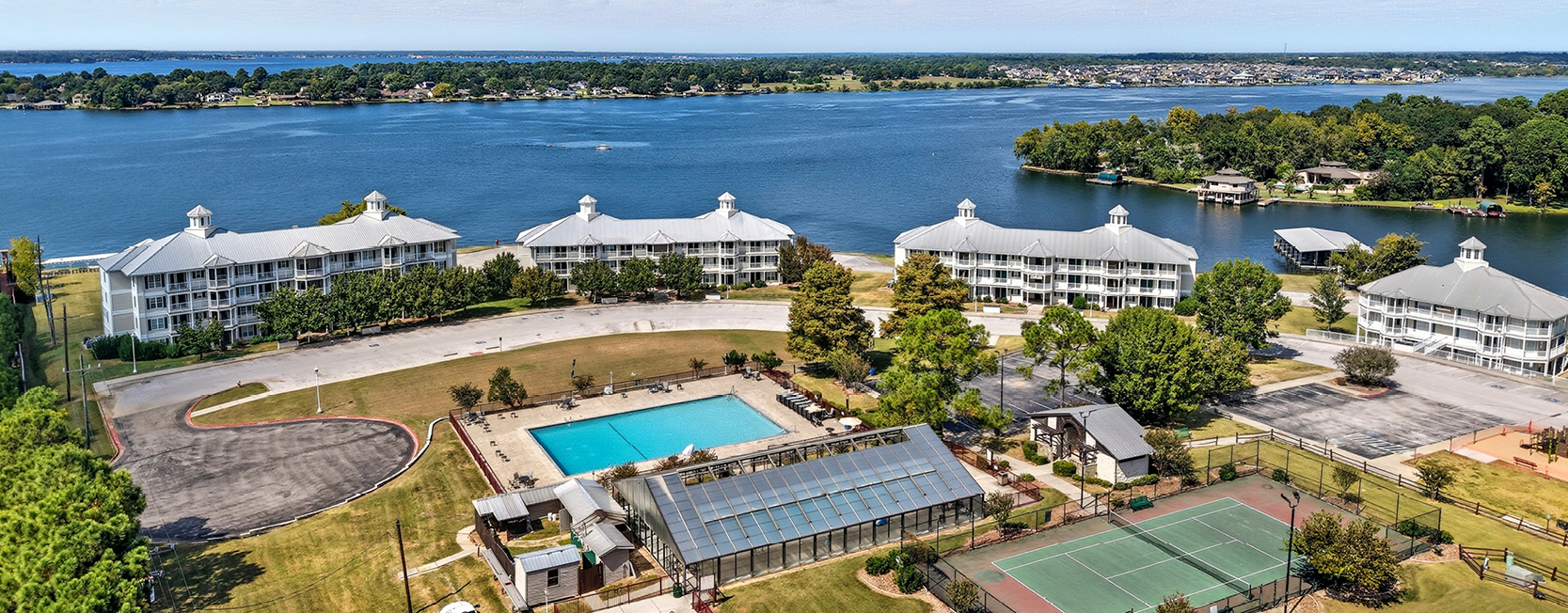 Aerial overview featuring apartment buildings, a swimming pool, tennis court, adn various other amenities with a lakeside view.