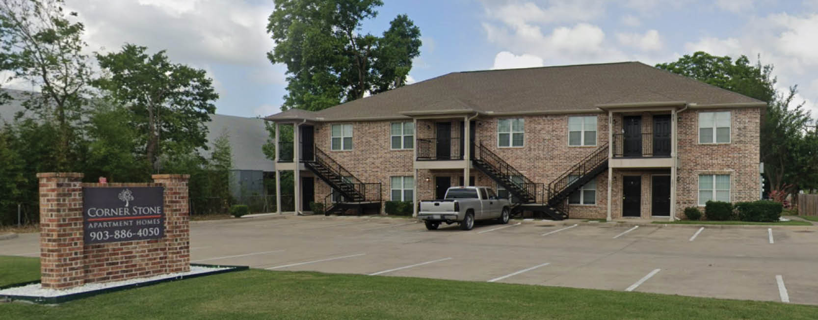 Two-story brick apartment building with exterior staircases and a mostly empty parking lot. A brick sign in the foreground reads “Corner Stone Apartment Homes” with a phone number, and trees surround the property under a partly cloudy sky.